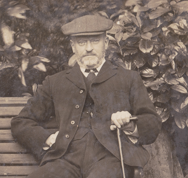 A sepia photo of a light skinned, bearded man in a dark suit and wool flat cap sitting on a bench and holding a walking stick in his left hand.